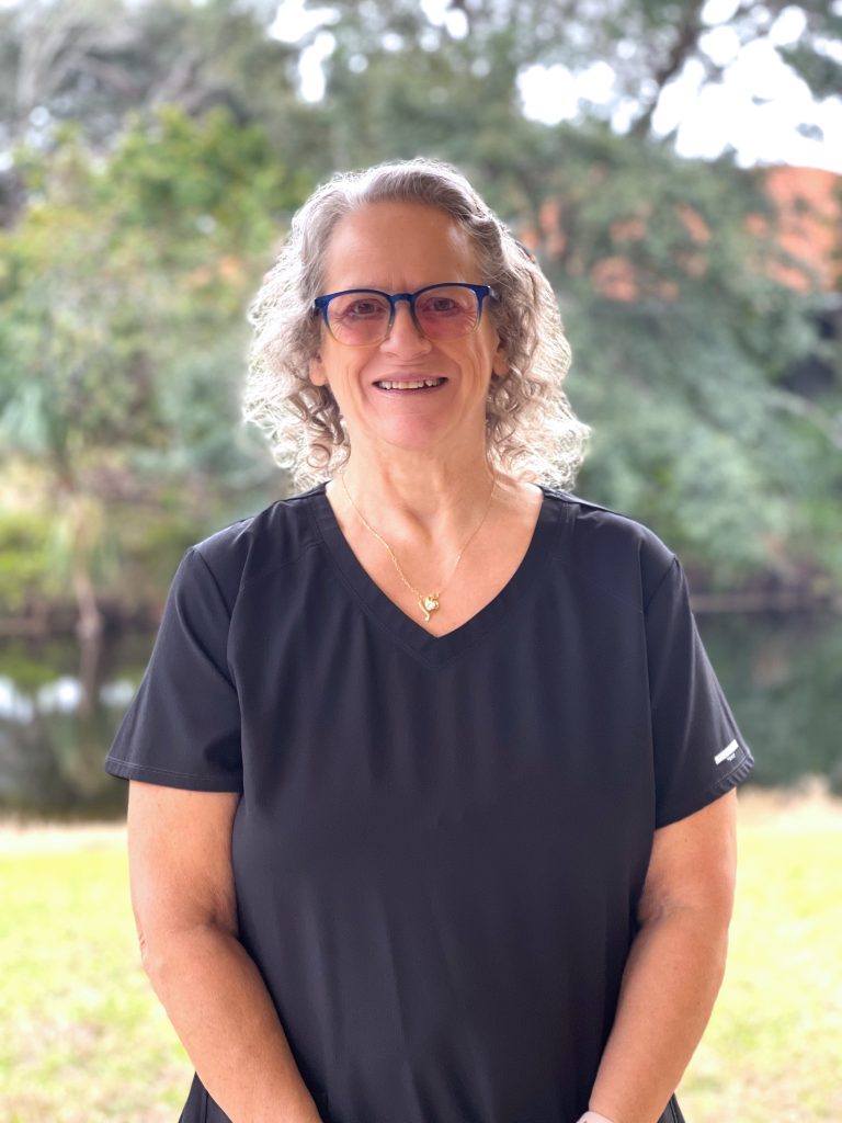 A smiling woman with curly hair, wearing a black top and glasses, stands outdoors by a body of water surrounded by green foliage.