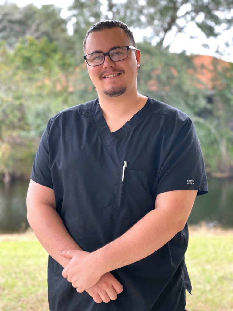 A smiling man in black scrubs stands outdoors near water, with trees in the background. He has glasses and a relaxed posture.