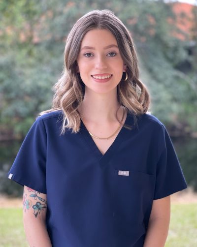 A young woman with shoulder-length hair smiles, wearing a navy scrub top, with visible tattoos and a natural outdoor background.
